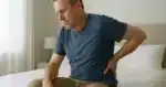 man sitting on bed and nursing his back in pain as a conceptual representation of the choice between consulting a chiropractor and orthopedic doctor